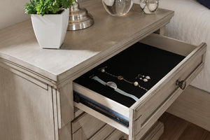 Mesa de noche de madera de color beige claro con un cajón abierto que muestra un organizador de joyas negro para guardar un reloj y joyas, en un ambiente de dormitorio.