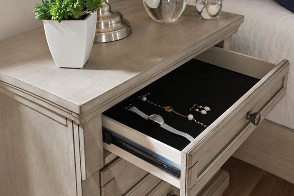 Mesa de noche de madera de color beige claro con un cajón abierto que muestra un organizador de joyas negro para guardar un reloj y joyas, en un ambiente de dormitorio.