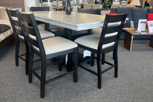 Juego de comedor alto con mesa cuadrada en acabado gris claro y cuatro sillas negras con asientos acolchados beige. El conjunto está exhibido en una tienda de muebles con decoración moderna al fondo.