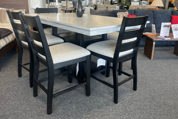 Juego de comedor alto con mesa cuadrada en acabado gris claro y cuatro sillas negras con asientos acolchados beige. El conjunto está exhibido en una tienda de muebles con decoración moderna al fondo.