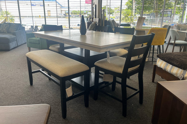 Juego de comedor alto con mesa cuadrada gris claro, banco acolchado beige y sillas negras de respaldo horizontal. El conjunto está exhibido en una tienda de muebles con ventanales amplios y decoración moderna.