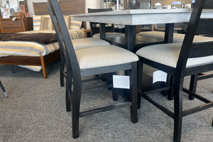 Primer plano de juego de comedor alto con mesa cuadrada gris claro y sillas negras con asientos acolchados beige. El conjunto está exhibido en una tienda de muebles junto a una cama y decoración contemporánea.