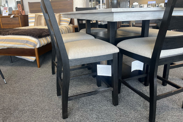 Primer plano de juego de comedor alto con mesa cuadrada gris claro y sillas negras con asientos acolchados beige. El conjunto está exhibido en una tienda de muebles junto a una cama y decoración contemporánea.
