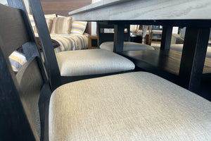 Detalle de sillas negras de comedor con asientos acolchados beige junto a mesa alta gris claro con repisa inferior. El conjunto está exhibido en una tienda de muebles con cama y decoración moderna al fondo.
