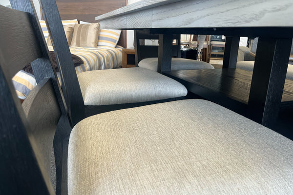 Detalle de sillas negras de comedor con asientos acolchados beige junto a mesa alta gris claro con repisa inferior. El conjunto está exhibido en una tienda de muebles con cama y decoración moderna al fondo.