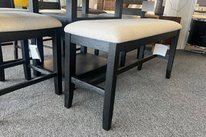 Banco alto para comedor en acabado negro con asiento acolchado beige y base reforzada. El banco forma parte de un juego de comedor moderno exhibido en una tienda de muebles sobre alfombra gris.