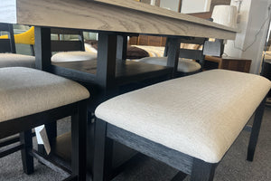 Banco alto para comedor con estructura negra y asiento acolchado beige, junto a mesa gris claro con repisa inferior. El conjunto moderno está exhibido en una tienda de muebles con decoración contemporánea.