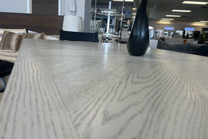 Primer plano de mesa alta de comedor en acabado gris claro con textura tipo madera y jarrón decorativo negro. El conjunto está exhibido en una tienda de muebles con camas y decoración moderna al fondo.
