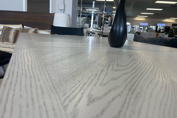 Primer plano de mesa alta de comedor en acabado gris claro con textura tipo madera y jarrón decorativo negro. El conjunto está exhibido en una tienda de muebles con camas y decoración moderna al fondo.