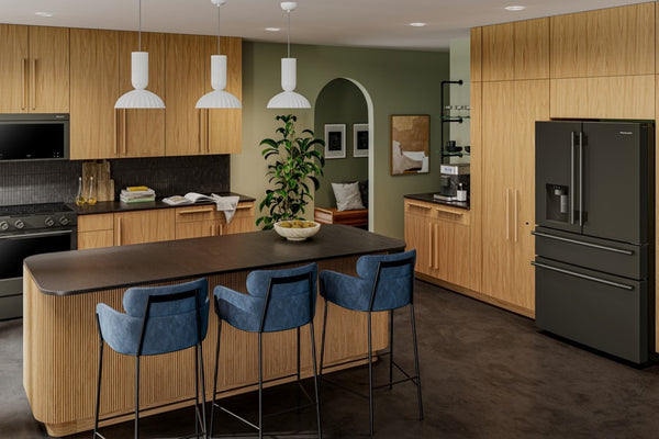 Un interior de cocina moderna con gabinetes de madera clara, una isla de madera oscura, tres taburetes de barra azules, lámparas colgantes, una pared verde y un refrigerador de puerta francesa de color gris oscuro.