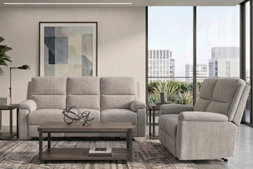 Juego de sala contemporáneo tapizado en tela gris claro, compuesto por sofá y loveseat reclinable, ambientado con mesa de centro, mesas auxiliares y grandes ventanales.