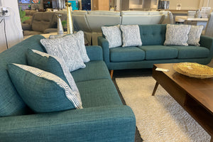 Juego de sala moderno con sofá y loveseat tapizados en tela azul, cojines decorativos con patrones geométricos en blanco, mesa de centro de madera oscura y alfombra clara en una sala de exposición.