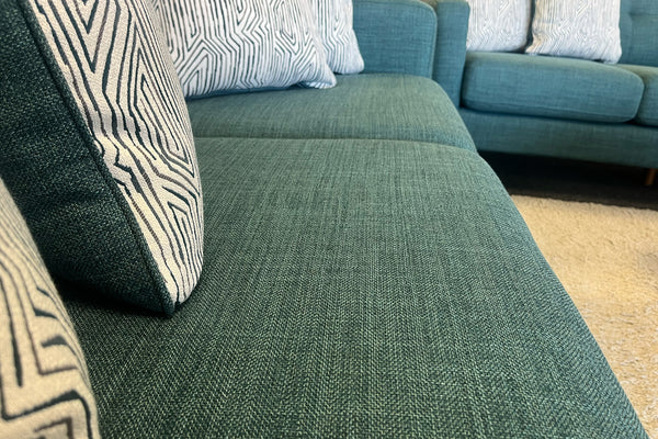 Primer plano de sofá tapizado en tela azul con textura tejida, acompañado de cojines decorativos con patrones geométricos en blanco y negro, en una sala de exhibición con alfombra clara.