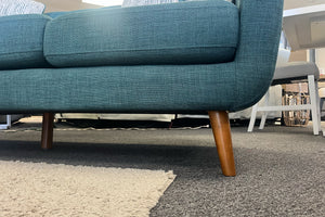 Primer plano de sofá moderno tapizado en tela azul con patas de madera inclinadas y cojines acolchados, ubicado sobre una alfombra beige en una sala de exhibición de muebles.