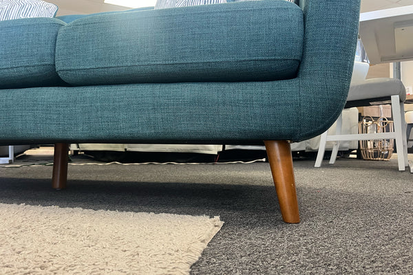 Primer plano de sofá moderno tapizado en tela azul con patas de madera inclinadas y cojines acolchados, ubicado sobre una alfombra beige en una sala de exhibición de muebles.
