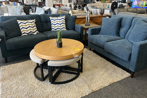 Juego de sala moderno con sofá y loveseat tapizados en tela azul oscuro, cojines decorativos con patrón geométrico en tonos beige, azul y blanco, mesa de centro redonda de madera con bancos integrados y alfombra beige en una tienda de muebles.