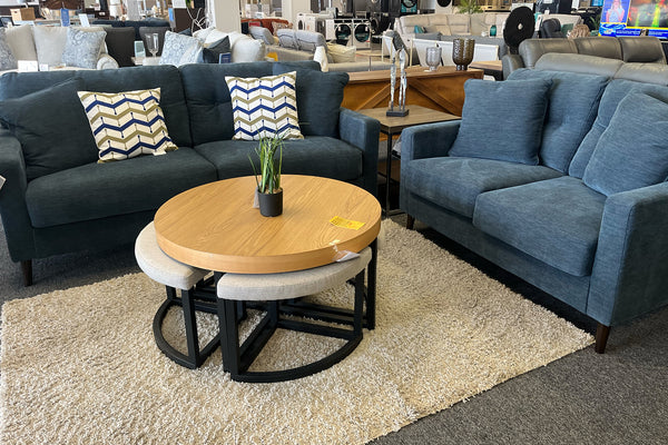 Juego de sala moderno con sofá y loveseat tapizados en tela azul oscuro, cojines decorativos con patrón geométrico en tonos beige, azul y blanco, mesa de centro redonda de madera con bancos integrados y alfombra beige en una tienda de muebles.