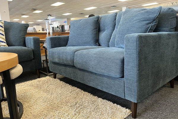 Loveseat moderno tapizado en tela azul oscuro con cojines acolchados a juego, patas de madera oscura y alfombra beige de textura suave en una sala de exhibición de muebles.