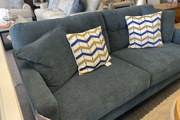 Sofá moderno tapizado en tela azul oscuro con cojines acolchados a juego, patas de madera oscura y alfombra beige de textura suave en una sala de exhibición de muebles.