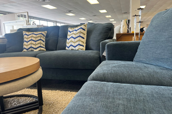 Sala de exhibición con sofá y loveseat tapizados en tela azul oscuro, cojines decorativos con patrón geométrico en tonos beige, azul y blanco, mesa de centro redonda de madera y alfombra beige de textura suave.