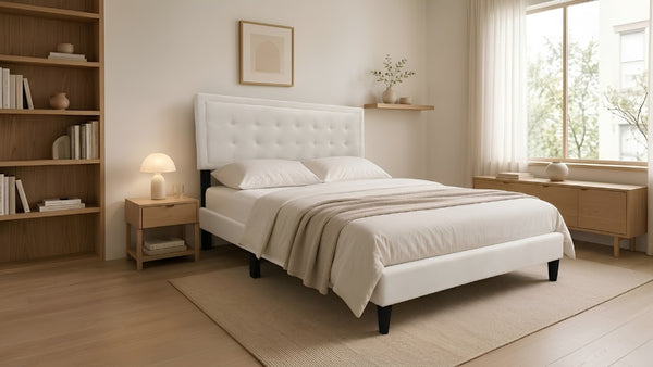 Cama Queen Kai con tapizado en tela color crema, cabecera capitonée de diseño rectangular y patas negras elevadas, en un dormitorio moderno con estantería y luz cálida.