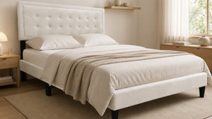 Cama Queen Kai tapizada en tela color crema con cabecera capitonée de diseño rectangular y patas negras elevadas, vista en un dormitorio moderno.