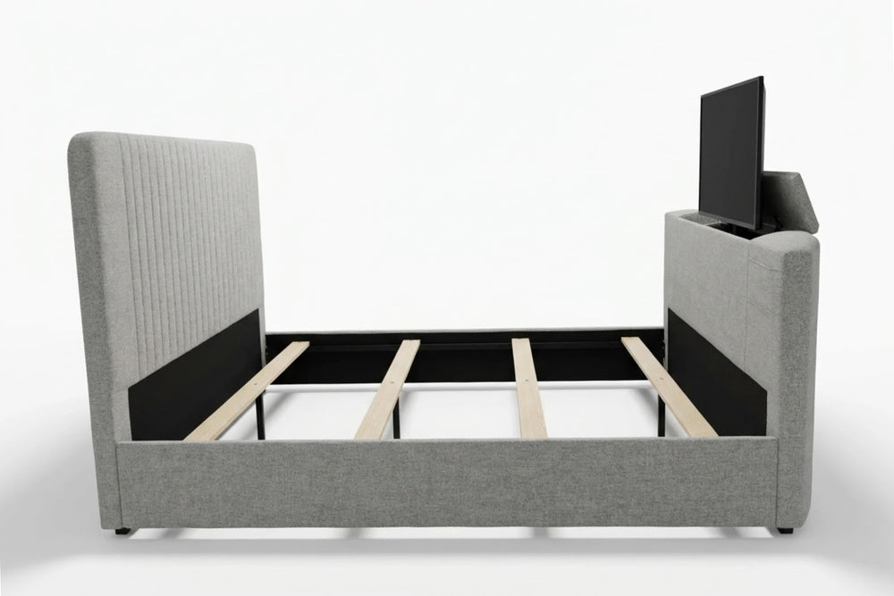 Cama moderna tapizada en tela gris claro, con cabecera alta acolchada y televisor integrado en el pie de cama. Presentada en dormitorio con ropa de cama blanca, mesas de noche de madera y decoración neutra.