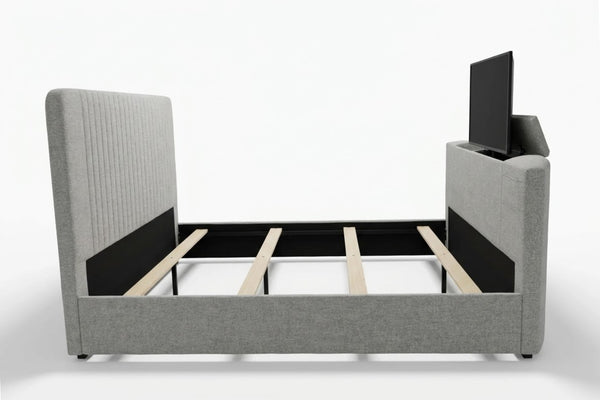 Cama tapizada en tela gris claro con cabecera alta de líneas verticales y estructura con soporte de madera. Incluye compartimento integrado en el pie de cama con televisor elevable.