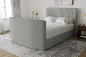 Una cama moderna tapizada con cabecero curvo y piecero a juego en tela gris claro, vestida con ropa de cama blanca y situada en un dormitorio luminoso con alfombra gris, suelo de madera y mesita de noche.