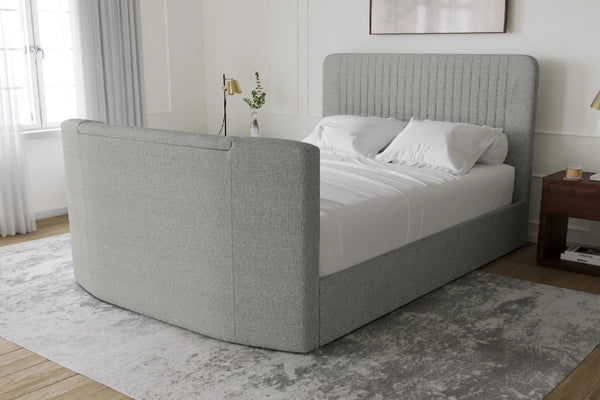 Una cama moderna tapizada con cabecero curvo y piecero a juego en tela gris claro, vestida con ropa de cama blanca y situada en un dormitorio luminoso con alfombra gris, suelo de madera y mesita de noche.