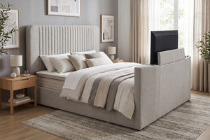 Cama moderna tapizada en tela gris claro, con cabecera alta acolchada y televisor integrado en el pie de cama. Presentada en dormitorio con ropa de cama blanca, mesas de noche de madera y decoración neutra.