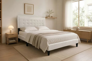 Cama Queen Kai con tapizado en tela color crema, cabecera capitonée de diseño rectangular y patas negras elevadas, en un dormitorio moderno con estantería y luz cálida.