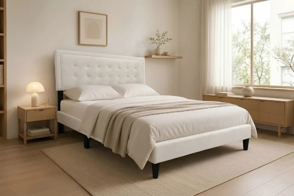 Cama Queen Kai con tapizado en tela color crema, cabecera capitonée de diseño rectangular y patas negras elevadas, en un dormitorio moderno con estantería y luz cálida.