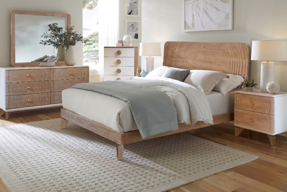 Un conjunto de muebles de dormitorio  que incluye una cama con cabecero de madera texturizada, una mesa de noche a juego y un gavetero con espejo, todo ello decorado con ropa de cama en tonos neutros y una lámpara sobre un fondo blanco.