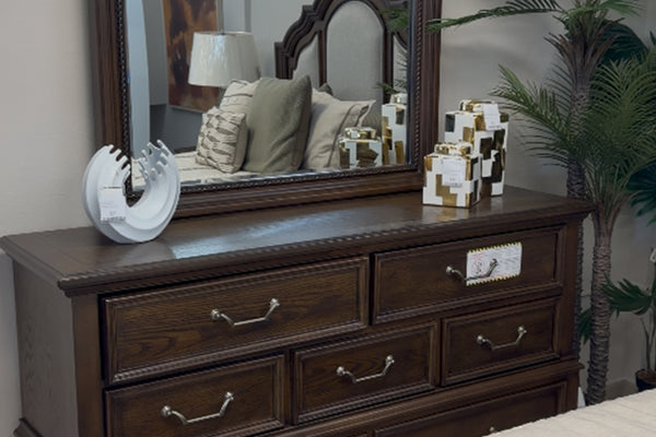 Gavetero hecho de madera marrón oscuro con espejo y con objetos decorativos. 
