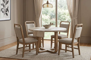 Juego de comedor de 5 piezas con mesa redonda de madera en acabado natural y cuatro sillas tapizadas en tela beige. Conjunto exhibido en un comedor luminoso con ventanal, cortinas claras y decoración de estilo contemporáneo.