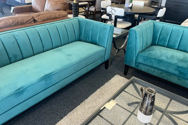 Juego de sala moderno con sofá y loveseat tapizados en terciopelo color verde, respaldo con costuras verticales decorativas, mesa de centro de vidrio y alfombra clara en una tienda de muebles.
