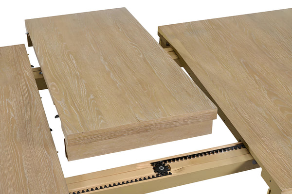 Un primer plano de una mesa de comedor de madera de color roble claro o blanquecino con acabado de vetas de madera, mostrando su mecanismo de extensión. Se ve una hoja de extensión central que está siendo insertada entre las dos mitades de la mesa. El mecanismo debajo incluye una guía metálica (posiblemente de color dorado pálido) y una cremallera dentada negra para deslizar las piezas.