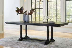 Una mesa de comedor rectangular de madera oscura, con patas torneadas y travesaños de refuerzo, situada en un comedor luminoso con una alfombra gris, floreros y copas de vino.