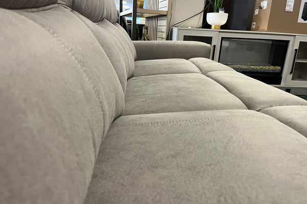 Primer plano de sofá reclinable tapizado en tela beige clara con asientos acolchados y costuras visibles, exhibido en una tienda de muebles junto a una consola decorativa con chimenea eléctrica.