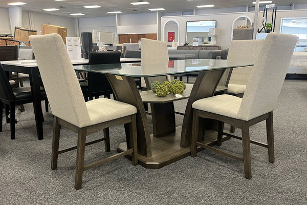 Juego de comedor contemporáneo con mesa rectangular de vidrio y base de madera oscura con estante inferior decorativo, acompañado de cuatro sillas tapizadas en color marfil. Exhibido en una sala de muebles con diseño elegante y funcional.