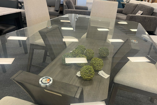 Vista superior de mesa de comedor con cubierta de vidrio templado y base de madera oscura con estante decorativo inferior. Incluye detalles decorativos y sillas tapizadas en color marfil, combinando funcionalidad y estilo contemporáneo.