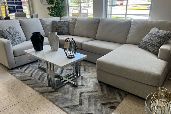 Sofá seccional moderno en tela gris claro con chaise longue a la derecha, cojines decorativos en tonos grises y mesa de centro blanca con base cromada. Exhibido sobre una alfombra geométrica gris en una sala de exhibición iluminada.