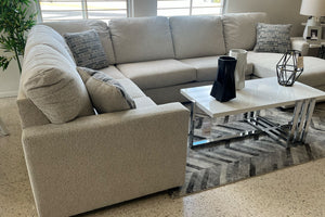 Vista lateral de sofá seccional en tela gris claro con cojines decorativos, chaise longue integrada y respaldo acolchado. Frente a una mesa de centro blanca con base cromada, sobre una alfombra geométrica gris en sala de exhibición.