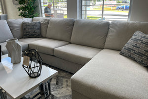 Sofá seccional contemporáneo en tela gris claro con chaise longue, cojines decorativos en tonos grises y asientos amplios. Ubicado junto a una mesa de centro blanca sobre una alfombra geométrica gris en una sala de exhibición iluminada.