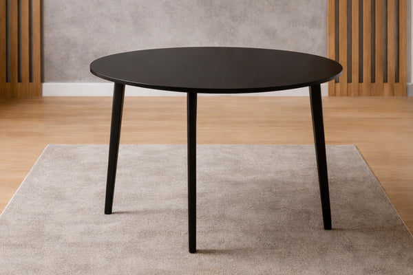 Mesa de comedor redonda negra con patas cónicas sobre una alfombra beige, piso de madera clara, estilo escandinavo minimalista.