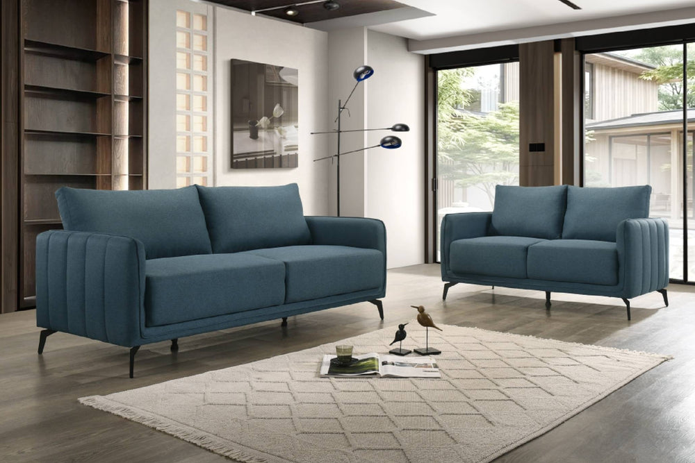 Juego de sala moderno en tela azul, compuesto por sofá y loveseat, con cojines amplios, brazos acolchados con líneas verticales y patas metálicas negras.