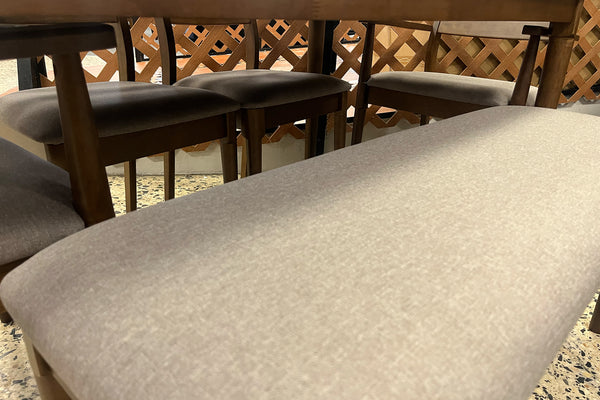 Primer plano de banco tapizado en tela gris claro junto a una mesa de comedor de madera oscura. Al fondo se observan sillas a juego con asientos acolchados, destacando la textura suave y el diseño contemporáneo del conjunto.