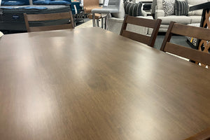 Vista cercana de mesa de comedor rectangular en acabado madera oscura con superficie amplia y lisa. Alrededor se observan sillas a juego de respaldo horizontal, exhibidas en una tienda de muebles con decoración contemporánea.