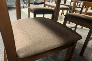 Primer plano de asiento tapizado en tela gris oscuro con textura suave, acompañado de sillas y mesa de comedor de madera en tono cálido. La imagen destaca los materiales, el acolchado y el acabado contemporáneo del conjunto.